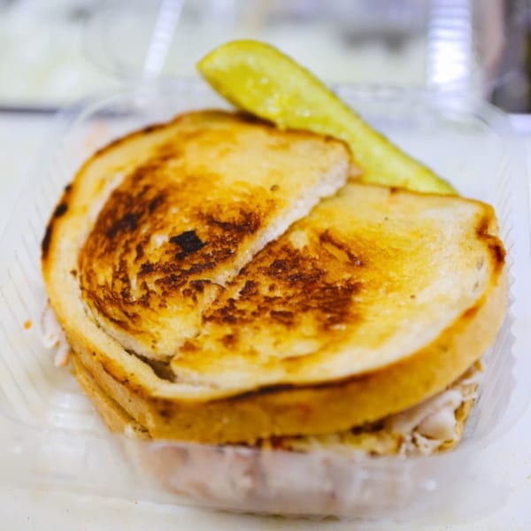 A grilled sandwich with toasted bread and a pickle spear in a clear plastic container.